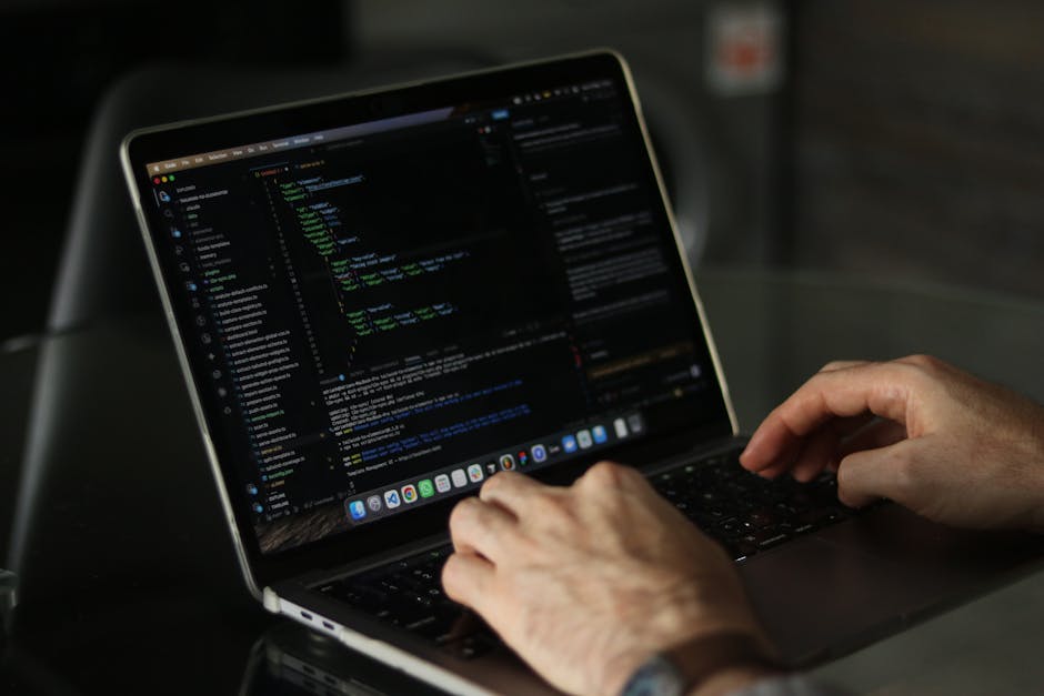 A close-up view of a person's hands typing on a MacBook laptop with a black keyboard, displaying a code editor with lines of programming code in various colors on the screen. The device is placed on a