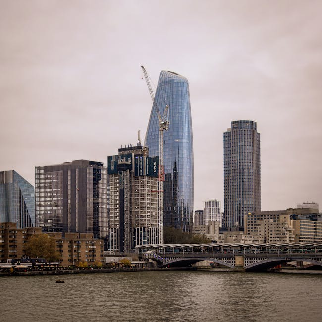 A view of urban riverfront with a mix of modern skyscrapers and construction sites, including a tall, curved glass building under construction with cranes positioned at the top. Surrounding buildings 